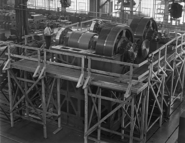 Speed increaser on the factory floor. This product was purchased by Allis Chalmers and used by the United States Corps of Engineers. Falk caption reads, "Low level dam site/generator drive. Large special speed increaser for Ozark dam site of Arkansas River... see Engineering File for exact details. This view shows partial assembly minus top covers and thrust bridge." Male employee in photograph is Don Ferch.
