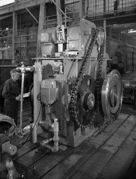 A male employee, Ed Grown, attends to a large ball mill drive. This product was purchased by Allis Chalmers and used by Pima Mining Company. Falk caption reads, "41 x 21.9Z1S speed reducer, ratio 5.308:1. 2 units on MO7-075276 and 4 identical units on MO 07-075220. One inching drive for all units furnished on MO7-067675. See Engineering File for all other exact details. Units include 230F H.S. coupling and 270F L.S. coupling."