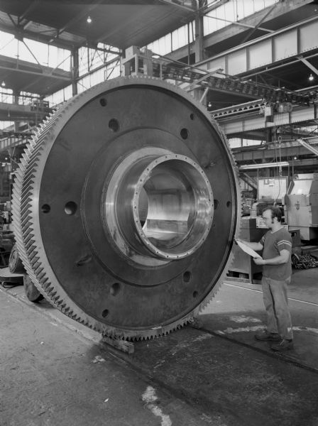 View of a 13 foot diameter hub gear that is a later component of a basic oxygen furnace drive. This gear was purchased and used by the Bethlehem Steel Corporation. Falk caption reads, "This gear was fabricated by Bethlehem Steel Corporation using a continuous forged ring for the rim section. They design this gear from the drawings of the original gear supplied with the Falk unit. The Falk Corporation cut the teeth and lapped this gear only." Male employee in photograph is Ronald Boettcher.