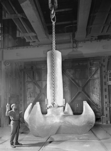 View of a large casting made in the foundry. A male employee, Arthur Beoman, stands next to the casting as it is being lifted. This unit was purchased by Clyde Iron Works. Original Falk caption reads, "32,000 pound large cast steel hook for lifting rig. See Engineering File for exact specifications."