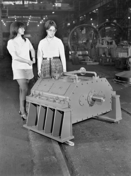 Two female employees, Virginia Breest on the left and Shirley Gunn on the right, attend to the speed increaser that is a later component of a jet engine compressor test stand. This unit was purchased and used by Avco-Lycoming. Falk caption reads, "Final assembly views of special 100,000 rpm dual path speed increaser... see model number 7-075305 in the Engineering File for exact details."