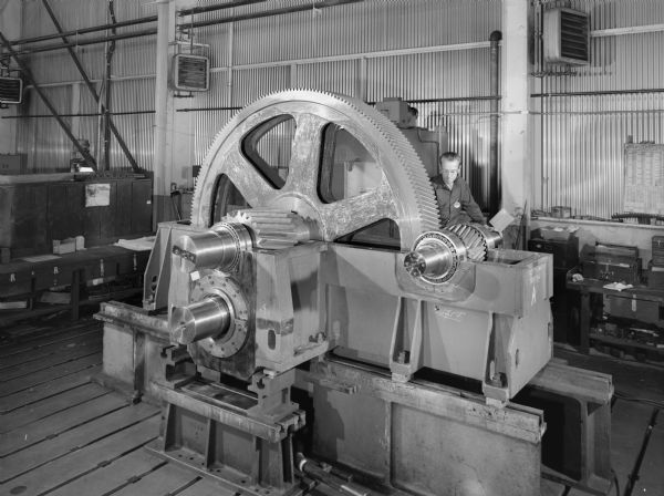 View of a combination reducer/pinion that is a component of a roughing mill for steel. This unit was purchased and used by the Eastern Carolina Steel Division. Original Falk caption reads, "This Falk drive replaces a special Swedish Morgardshammar reducer and pinion stand combination drive. It is the roughing mill drive in a steel plant and will accept billets 4 to 4 1/2 inches square. The roughing mill is the primary piece of equipment in this facility and its importance dictated a Falk drive. This particular mill is unusual in that the roll stand moves vertically for each of the 7 passes. The table and the drive are stationary. The 2.75 service factor is required to withstand the high loads imparted through the use of two flywheels to be installed on the high speed shaft. Design load: 4235 HP; Service load: 1540HP; Speed; 890 RPM input-121.75 RPM output; ratio: 7.310:1; Service factor; 2.75; Service: Heavy Duty; Duty: Intermittent." Male employee in photograph is Don Ferch.