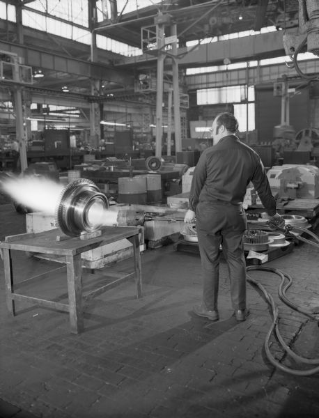 Original Falk caption reads, "This photo taken to illustrate pre-heating a large coupling hub by torch prior to making a shrink-fit mounting on a shaft." Male employee in photograph is Frank Alioto.