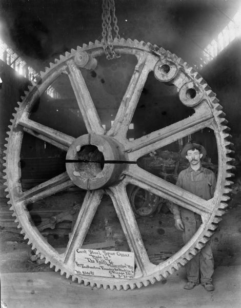 An unidentified Falk employee stands next to a large Steel Spur Gear. The sign in the photograph reads: "Cast Steel Spur Gear. Made by The Falk Co. Milwaukee, Wis. For Featherstone Foundry and Machine Co. of Chicago, Ills. Wt. 9340 lbs." This photograph appeared in the April 1974 issue of the "Falk Reflector".