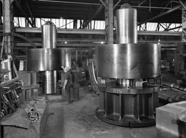 View of machinery castings that were a later component of a cement mill support roller. This unit was purchased by the Fuller Company and and used by Huron Cement. Original Falk caption reads, "Used on 17 foot x 19 foot 6 inch x 500 foot kiln. 90 inch x 41 inch face cast roller-pattern number 728-73-3-1143-90; weight-60,000 pound roller only; shaft forged-27 inch diameter x 11 foot 4 inches long. 26,000 pound shaft only. Total weight of assembly-86,000 pounds." Male employee in photograph is Joseph J. Rozman.