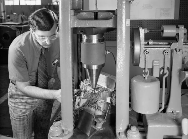 A male employee, John Lisiecki, attends to a test of a worm gear. Original Falk caption reads, "This is a close-up of the tooth model in the Tinius-Olsen tester for project 75E-27. Point loads are applied through a one inch diameter ball in a loading cone which is attached to a 50,000 pound capacity load cell."