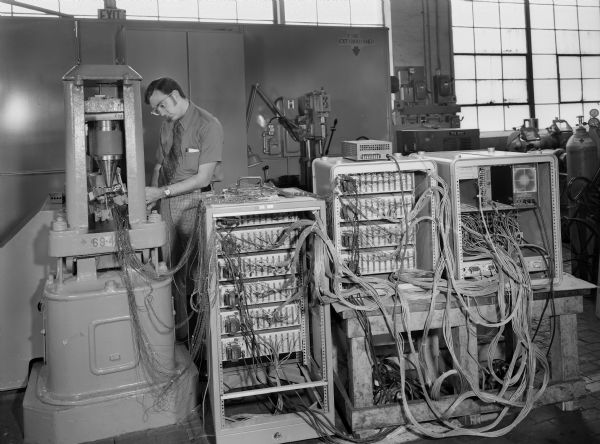 A male employee, John Lisiecki, attends to a research test on a worm gear tooth model. Original Falk caption reads, "Overall view of test setup for project 75E-27. This view shows the back of the strain measuring and recording instrumentation which highlights the wiring which connects the instrumentation to the strain gauges on the tooth model."