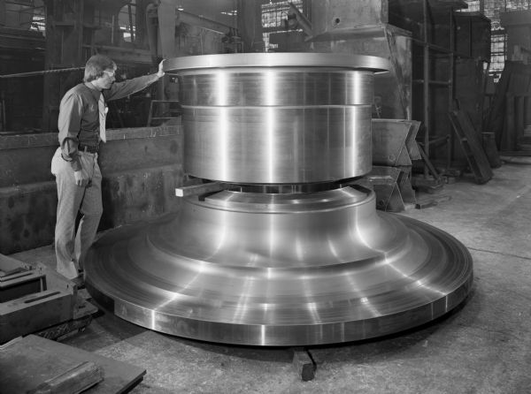 William Rhody, a Falk employee, inspects a 2 piece trunnion. This particular unit was purchased by F.L. Smidth. Original Falk caption reads, "Trunnion-two piece design: both pieces are rough machined. The pieces are separated by two by fours showing the weld scarf before preparations are made for welding the two pieces together. Trunnions hubs: approximate rough weight 16,000 pounds, pattern number DXL-291-1, material ASTM A-27 Gr. 60-30 annealed. Trunnion Flange: approximate rough weight 28,000 pounds, pattern number DXL-291."