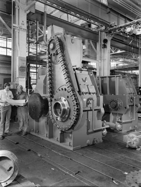 Duane Hanson and Norbert Winski attend to a large reducer and incher. This unit was purchased by Allis Chalmers and used by the Cleveland Cliffs Iron Company-Tilden II Expansion. Original Falk caption reads, "Rockcyl mill main reducer and incher. Main reducers for dual pinion mill drive with 130 F unit incher. Chain and sprocket connection between incher and main with a shift-able drive sprocket hub disconnect feature."