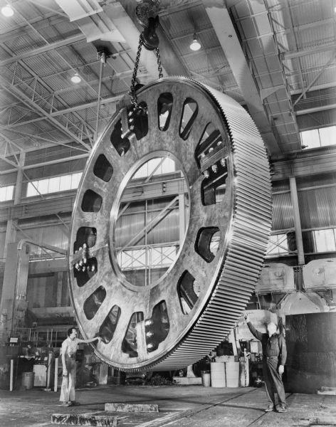 A large single helical ring gear hangs from the ceiling in the Falk factory. This later component of a kiln drive was purchased by Allis Chalmers and used by the Cleveland Cliffs Iron Company-Tilden II Mine. Original Falk caption reads, "EG 50127 copied by Brackett on April 4, 1979 from retouching 1600-P30. 1600P30 retouching made from color transparency 275TF-2870." Male employees in photograph are Joe Cocroft and Theodore Lutzen.