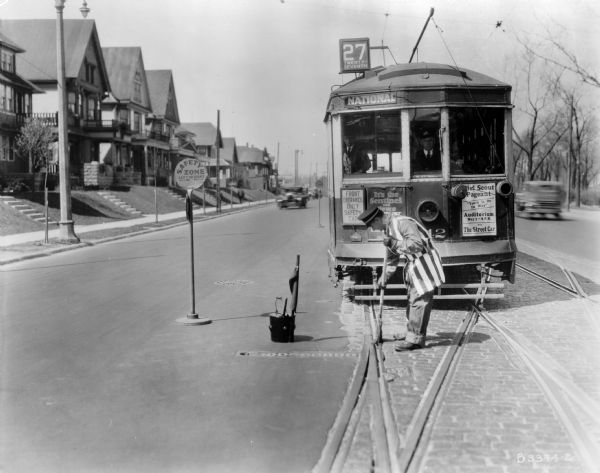 A street cleaner sweeps the tracks in a residential neighborhood, as the streetcar and its operator and passengers wait. Original Falk caption reads, "Photos borrowed from Milwaukee County Transport for use in brochure 850801. Originals returned to County. Milwaukee streetcars."