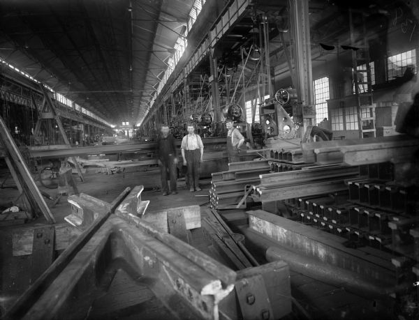 Three Falk employees stand inside of the Falk factory. Around them are various steel beams, machines, and tools. Original Falk caption reads, "Centennial historical photos of old negative which could be lost forever. Probable dates prior to World War One."