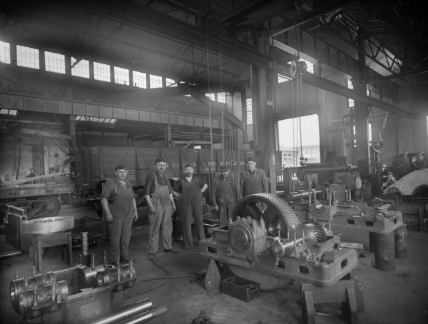 Five Falk employees stand inside the foundry. They stand among various gears and machinery. In the background, two more employees are visible. Original Falk caption reads, "Centennial historical photos of old negative which could be lost forever. Probable dates prior to World War One."