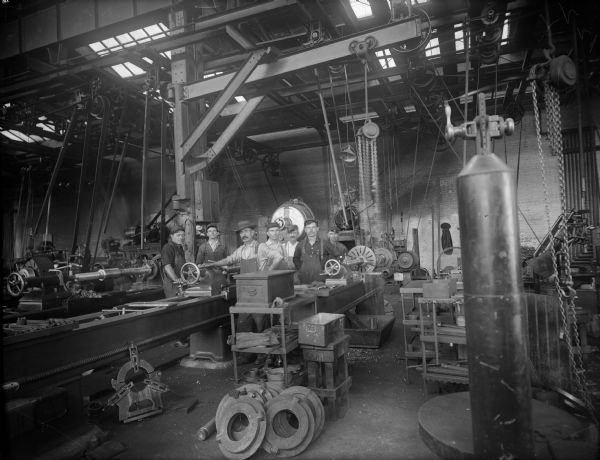 Six Falk employees stand in machine shop. There are numerous machines and gear implements surrounding them. Original Falk caption reads, "Centennial historical photos of old negative which could be lost forever. Probable dates prior to World War One."