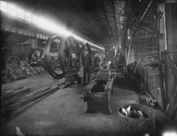 Two men stand near several large gear castings and numerous machines. Original Falk caption reads, "Centennial historical photos of old negative which could be lost forever. Probable dates prior to World War One."