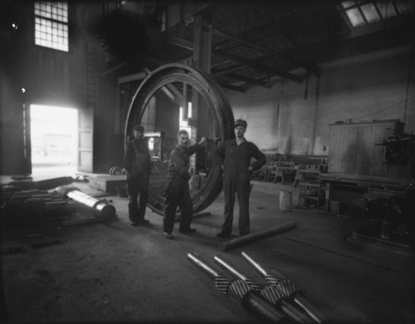 Three Falk employees stand with a large gear in the foundry. Original Falk caption reads, "Centennial historical photos of old negative which could be lost forever. Probable dates prior to World War One."