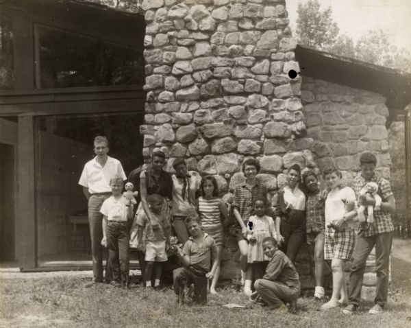 Group portrait of children and adults, some carrying stuffed animals, in front of the library at the Koinonia Children's Camp. Ruthie Hartford in the center in a striped shirt.