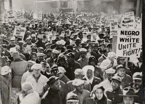 Shell labor rally at an unknown location.  Signs include the sayings, "Equal Pay for Women," "Negro and White, Unite and Fight," and "Let the Packers Pay Our Increase in Taxes!."