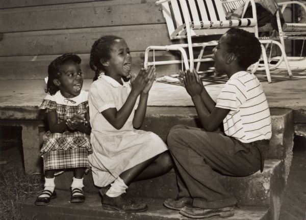 Three children sitting on a porch singing together and playing pattycake.