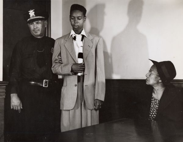 Unidentified image of a young man in a suit and tie being escorted by a police officer into a court room.