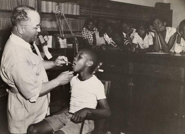 A boy receiving dental work at the Highlander Folk School.  A group of children look on from over a counter.