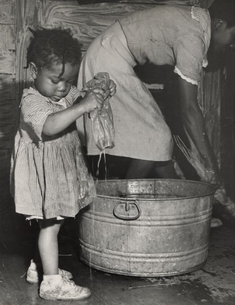 A young girl helps her mother with laundry by wringing out a pair of shorts next to a wash basin.
