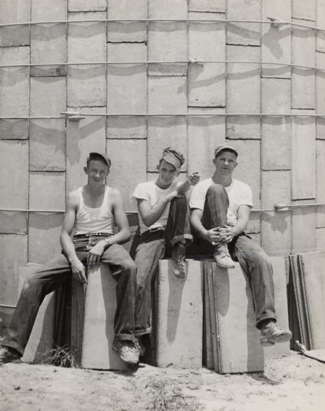 Private building of Farmers Union Fertilizer Co-op, with three men in work clothes sitting in front. Junior Marlow on right.