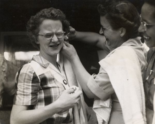 A smiling woman with a towel is swabbing the side of a woman's head at a CIO or Amalgamated Transit Union workshop at Highlander Folk School. Another woman looks on.