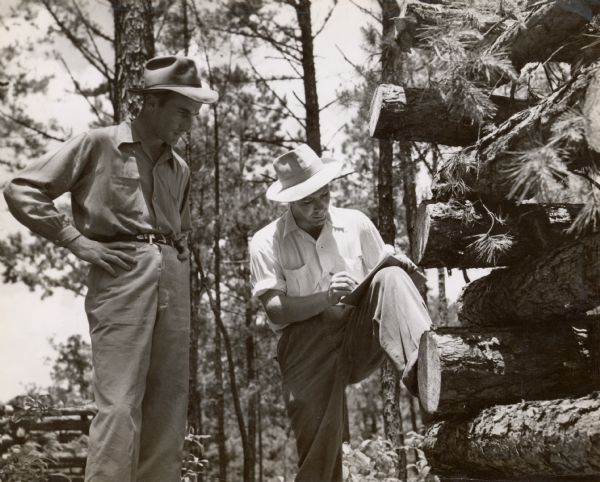 Two men wearing fedora hats are standing by stacked logs. One of the men is writing in a notebook.