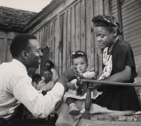 An African American man, possibly Medger Evers, and woman, with a baby at Highlander Folk School. A group of women can be seen in the background.