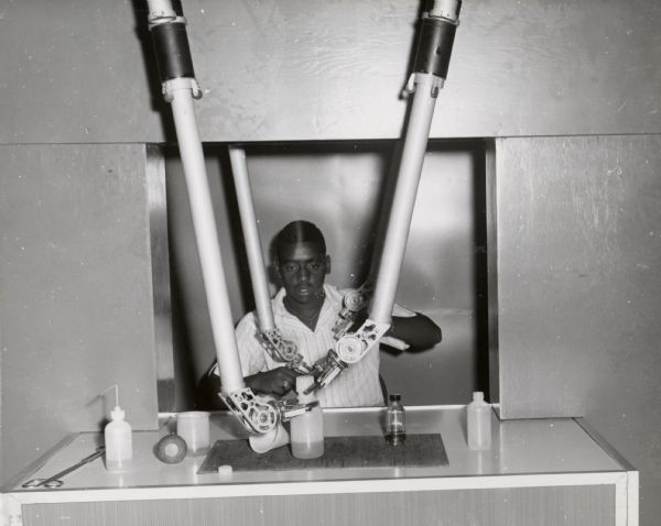 An African American man operating a series of large, mechanical arms to manipulate various bottles and scientific instruments. At the Oak Ridge Atomic Museum.