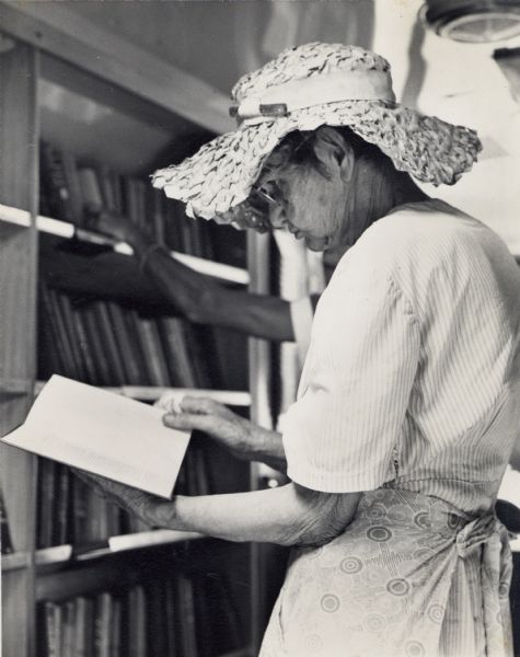 An elderly woman in a straw sun hat reading a book in the Highlander Folk School Library.