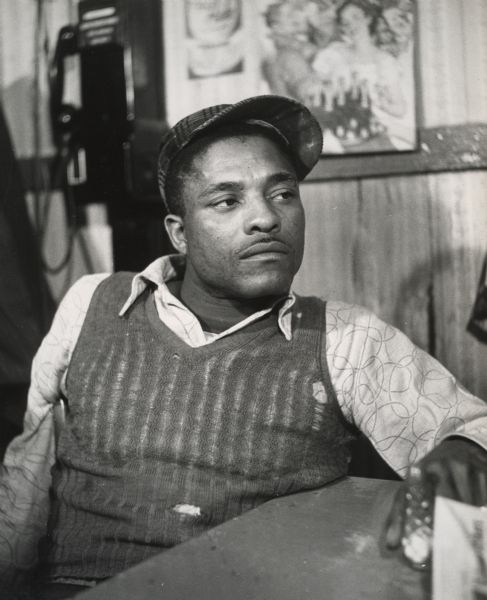 An unidentified African American man, wearing a torn sweater and a corduroy baseball cap, sitting at a table.