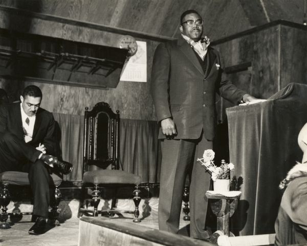 An African American man, perhaps a clergy member, wearing a suit and scarf, standing in front of an altar. Another man is sitting behind him. Possibly Leroy Brown on the left.