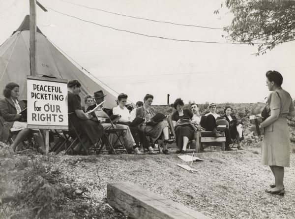 Zilphia Horton playing an accordion and singing on a picket line. A group of men and women sit in chairs in front of a tent. Sign on left reads, "Peaceful Picketing for Our Rights." Bill Elkuss, third from right, seated; Mary Elkuss, fourth from right, seated.