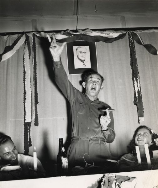 A man holding a cigar is gesturing wildly.  A portrait of Franklin Delano Roosevelt hangs behind him.  An unidentified man and Catherine Winston sit on either side of him. Part of an event at Highlander Folk School.