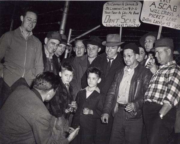 Textile workers strike in Alabama.  Signs in back read, "This Company Is Unfair To The Laboring Class + It Is Your Fight As Well As Ours.  Don't Scab.  Dallas Local #43 TWUA CIO" and "Only a Scab Will Cross a Picket Line. Dallas Local #43 TWUA CIO." Two young boys are surrounded by men listening to a man with his back to the camera.