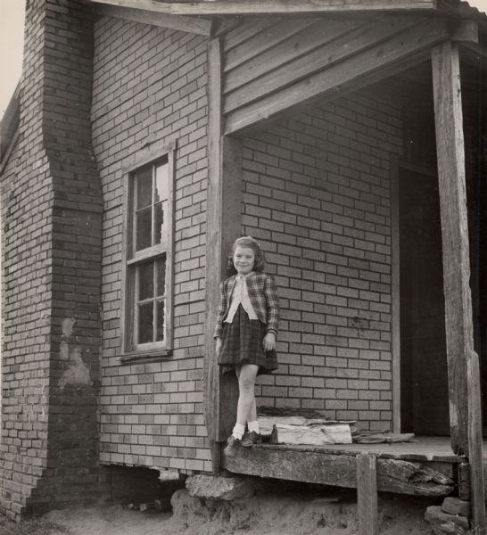 A girl in a plaid skirt and jacket, posing on a porch.