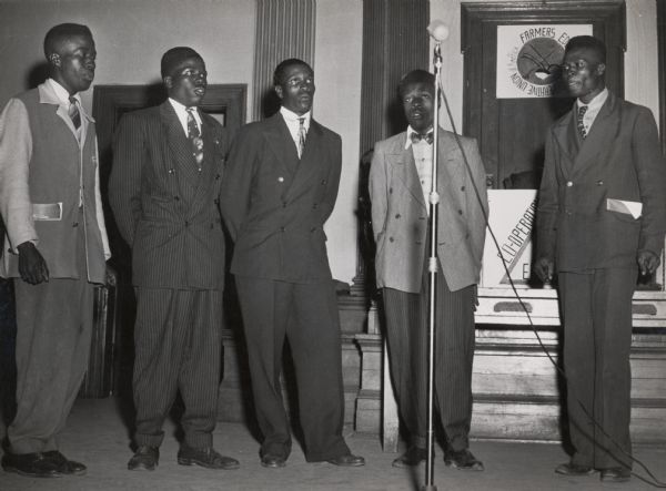 Five African American men wearing suits and singing into a microphone.  There is a small farmers union sign on the back wall.