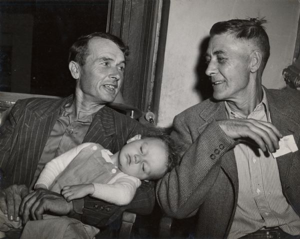 Two men sitting, one holding a baby in his arms, attending a Farmers Union Workshop.