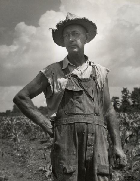 An unidentified farmer wearing overalls and a straw hat standing in a field.