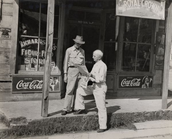 Two men reading a newspaper outside of a storefront.  The store's sign, partially obstructed, reads, "Richards[on]'s Flour & Feed[?] Stor[e].  Coca-cola, Royal Crown Cola, Pepsi Cola, and Red Seal Snuff advertisements adorn the outside.
