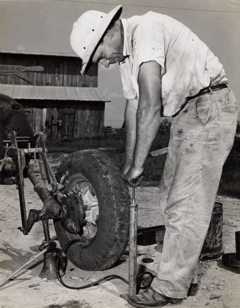 A man with a safari style hat pumping air into a tire of a farming apparatus.
