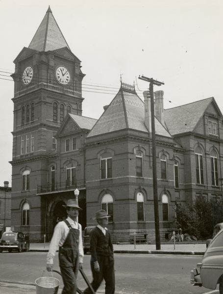 Two men wearing hats, one of whom is carrying a bucket and a tool, walking across the street from a large building.