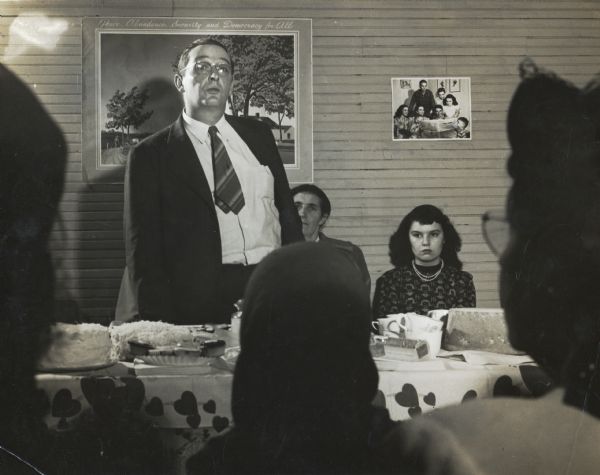 A man with a short tie standing up during an Alabama Farmers Union Meeting.  An image in the background reads, "Peace, Abundance, Security, and Democracy for ALL."