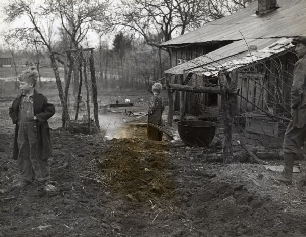 A man and two boys standing near a shack with large cauldron in front, looking in different directions.  They are neighbors of Highlander Folk School.