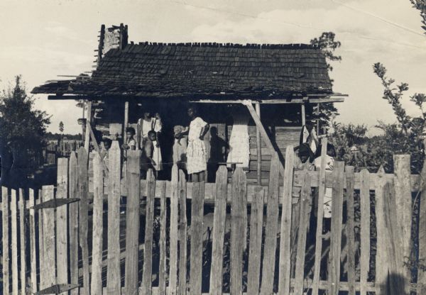 An African American family standing on the porch of their home, behind a wooden picket fence.