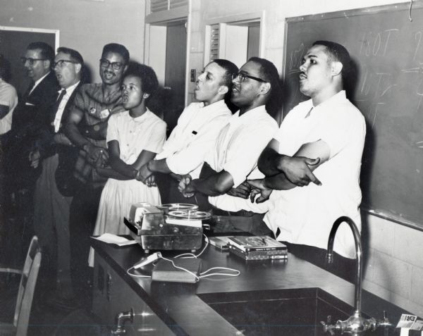 Highlander Folk School students and directors standing in a line, possibly singing together, at the San Padeo workshop. Third from left, Myles Horton, William Miller, Joyce Ladner, Dave Dennis, Bob Moses and Lawrence Guyot.
