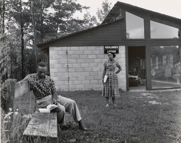 A man is reading a book on a bench, while a woman is standing in front of the entrance to the Highlander Library. A small group of people can be seen inside of the library.