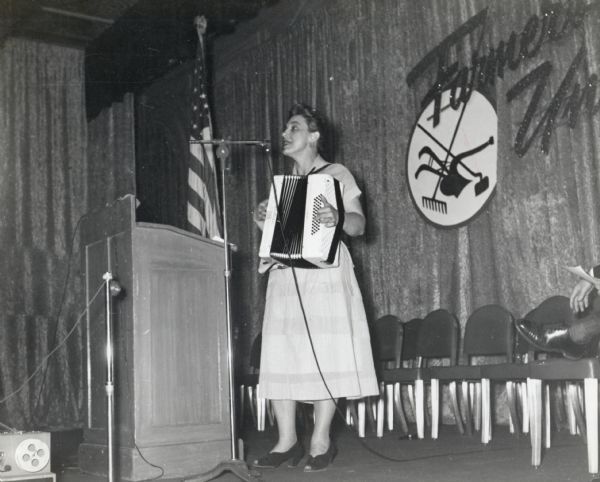 Zilphia Horton playing her accordion at a meeting of the Food and Tobacco Workers Union. On stage behind her is a "Farmers Union" sign with a graphic logo.